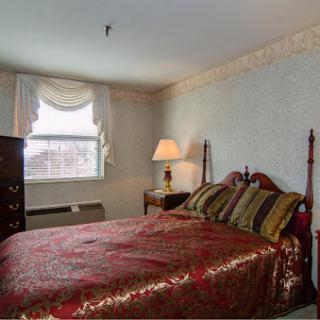 A cozy bedroom featuring a bed with decorative pillows, a lamp beside it, and a window letting in natural light.