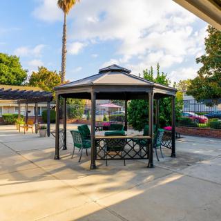 A cozy outdoor gazebo with seating, set in a sunny patio area with surrounding greenery and a palm tree.