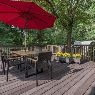 A cozy patio deck with a table and chairs under a red umbrella surrounded by greenery.