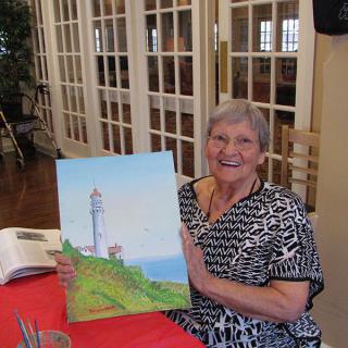 A woman proudly displays her painting of a lighthouse inside a room.
