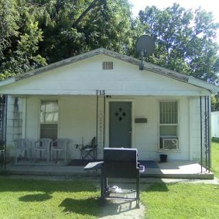 A quaint single-story house with a front porch and a yard surrounded by greenery.