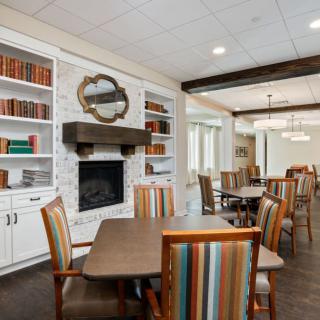 A cozy library and dining area with bookshelves, a fireplace, and colorful chairs around wooden tables.