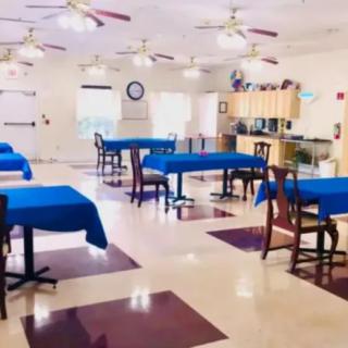 A spacious dining area with tables covered in blue tablecloths, featuring ceiling fans and a kitchen area in the background.