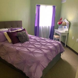 A cozy bedroom with purple bedding and green walls, featuring a window with light streaming in and a vanity table by the side.
