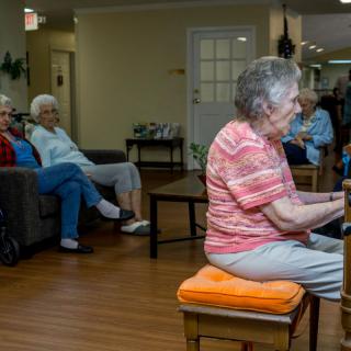 An elderly woman playing the piano in a communal area of a nursing home, with other senior citizens seated and listening.