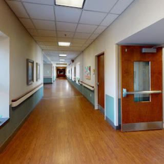 A spacious and well-lit hallway in a healthcare facility, featuring wooden flooring and closed doors along the sides.