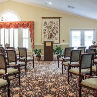 An interior view of a room set up with rows of chairs facing a podium, featuring decorative plants and a patterned carpet.