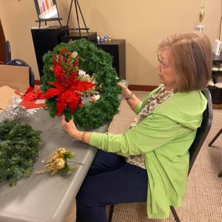 A woman decorates a wreath with festive elements at a table indoors.