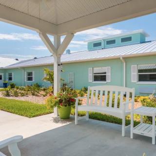 A serene outdoor area featuring a white bench under a covered porch, with pastel-colored buildings and a garden in the background.