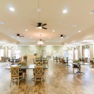 A spacious dining hall with neatly arranged tables and chairs, featuring wooden flooring and ceiling fans.