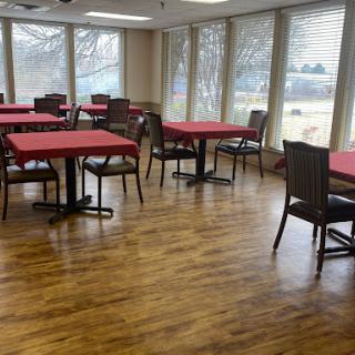 A spacious dining area with red tablecloths, featuring tables and chairs arranged for guests, illuminated by natural light from large windows.