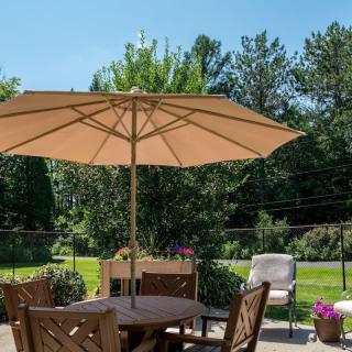 A sunny outdoor patio with a wooden table, chairs, and an umbrella, surrounded by lush greenery.