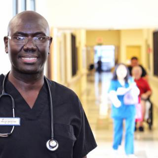 A smiling doctor in a hospital corridor with colleagues and patients in the background.