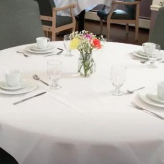 A neatly set round dining table with white tablecloth and a small floral centerpiece.