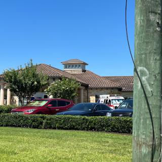 A view of a commercial building with parked cars in front, surrounded by lush greenery under a clear blue sky.