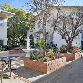 A peaceful residential courtyard featuring a central fountain, surrounded by brick planters with flowers and outdoor seating.