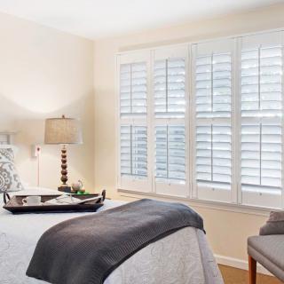 A cozy and well-lit bedroom featuring a bed with a tray, a bedside lamp, and large windows with shutters.