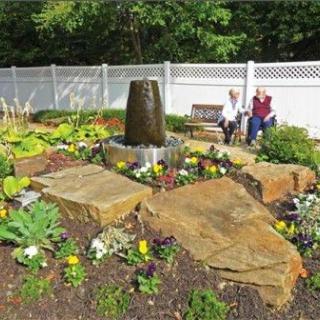 A beautifully landscaped garden with a central fountain, surrounded by flowers and large rocks. Two people are sitting on benches in the background, enjoying the serene setting enclosed by a white fence.