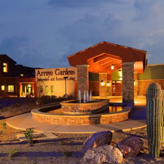 A well-lit entrance to a building with a fountain and desert landscaping at dusk.