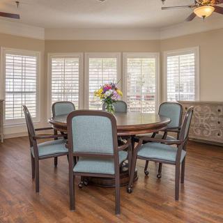 A cozy dining area with a wooden table and chairs, surrounded by windows with shutters and decorated with a vase of flowers.