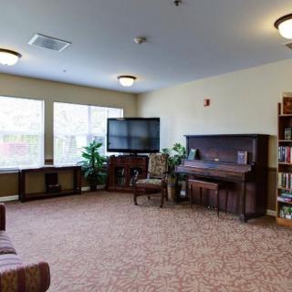 A cozy living room featuring a piano, TV, and bookshelves, with ample natural light from large windows.