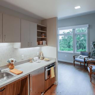 A modern kitchen with wood cabinets and bright lighting opens to a cozy living area with large windows.