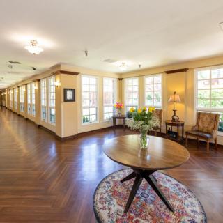 A spacious hallway with wooden floors, featuring a central round table and chairs alongside large windows.