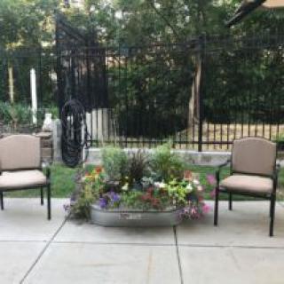 An inviting patio setup with two chairs and a planter filled with colorful flowers, surrounded by greenery.
