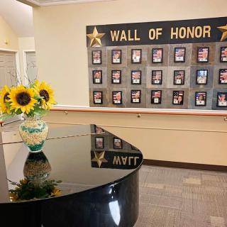 An interior space featuring a 'Wall of Honor' adorned with plaques and a grand piano topped with a vase of sunflowers.