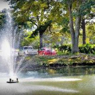 A scenic view of a park with a pond and fountain, surrounded by lush trees and parked cars.