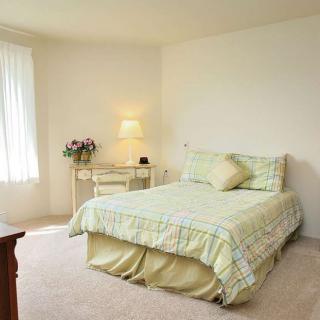 A cozy bedroom with a neatly made bed, nightstand with lamp, and a window letting in natural light.