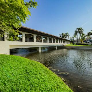 A scenic view of a bridge over a pond surrounded by lush greenery and trees under a clear blue sky.