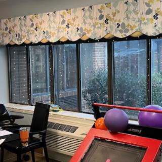 A cozy room with large windows, decorated with floral curtains, featuring a table and chairs along with exercise balls.