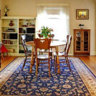 A cozy dining room with a wooden table and chairs, set on an ornate blue rug, surrounded by a bookshelf and a cabinet under a sunny window.
