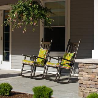 A cozy porch with two rocking chairs adorned with green cushions, surrounded by plants and a hanging basket.