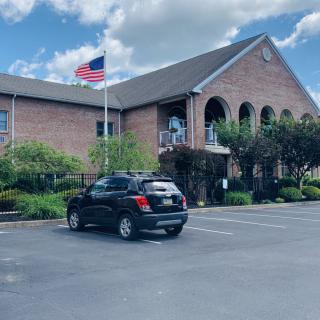A brick building with an American flag flying outside, surrounded by a parking lot and trees.