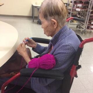An elderly person in a wheelchair knitting with pink yarn.