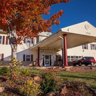A building entrance with a red car parked outside, surrounded by autumn foliage and clear blue sky.