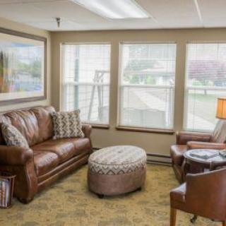 A cozy living room with a leather sofa, an armchair, and a patterned ottoman, featuring large windows and a warm atmosphere.