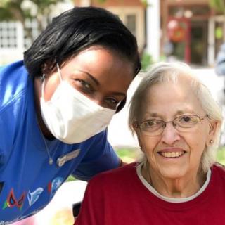 A caregiver wearing a mask posing with an elderly person outdoors, both smiling.