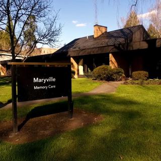 A memory care facility with a sign in front, surrounded by green grass and trees under a clear sky.