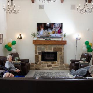 A cozy living room with a stone fireplace, decorated with balloons and a television mounted above. Two people are seated in the space.