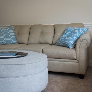 A cozy living room setup featuring a beige sofa with blue patterned cushions and a round ottoman.