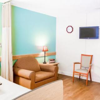 A well-lit hospital room with a bed, armchair, wooden chair, and a wall-mounted television.