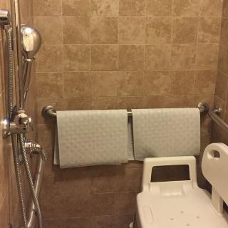 A shower area with tile walls, equipped with a shower chair, handrails, and two towels hanging on the wall.
