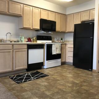 A modern kitchen with light wooden cabinets, black and white appliances, and tiled flooring.