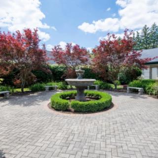 A beautifully landscaped garden featuring a central fountain surrounded by trees and a brick pathway.