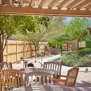A cozy backyard patio with wooden chairs and table set under a pergola, surrounded by lush greenery and a well-maintained garden.