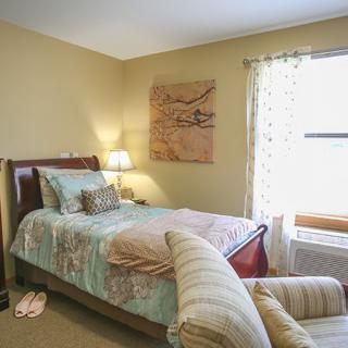 A cozy bedroom with a neatly made bed, soft lighting from a bedside lamp, and natural light streaming through the window.