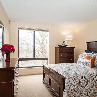 A cozy bedroom with wooden furniture and a large window, featuring a neatly made bed and decorative elements.
